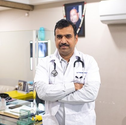 Dr. Prakash Joshi, a man in a white lab coat with a stethoscope around his neck, standing with arms crossed in a medical office.