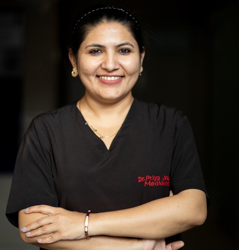 Dr. Priya Joshi, a smiling woman in medical scrubs with "Dr. Priya Joshi Medident Clinic" embroidered on her shirt.